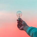 A hand holding a clear light bulb against a gradient blue and pink sky, symbolizing insight, influence, leadership dynamics, and power.
