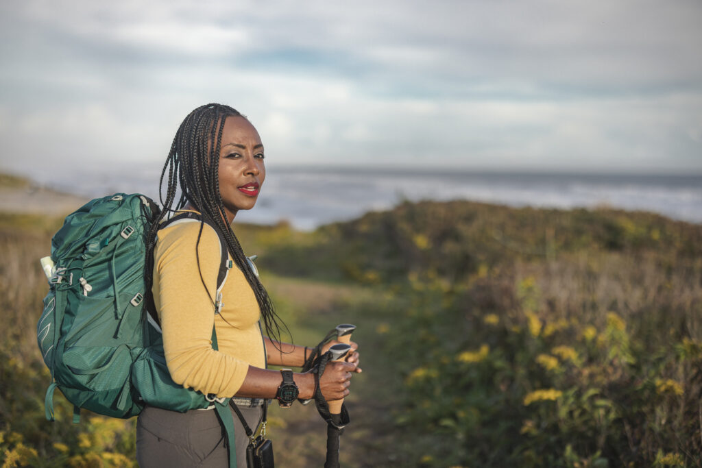 African American woman wearing a backpack while walking a hiking trail and carrying hiking poles with a perturbed facial expression. group communication and unspoken expectations