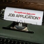 A vintage green typewriter sits on a dark wooden desk with a sheet of paper loaded into its roller. The paper displays the words "Do applicants hate your JOB APPLICATION" typed in bold, all-caps lettering, creating a classic and focused visual that evokes themes of employment, retro technology, and formal documentation. The focus of the blog is job application privacy.